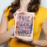 This image features artist JAK, a vibrant and expressive individual, holding a small, rectangular art piece with the bold text "Keep F***ing Going" painted in a striking, abstract style. The background hints at a casual setting with the woman dressed in a bright yellow top and white trousers, accessorized with a pop of red lipstick.